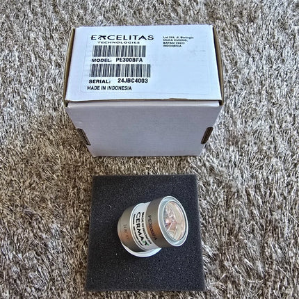 A small cylindrical metal component labeled 'EXCELITAS' sits on a black foam insert, with its branded cardboard box placed behind it on a shaggy gray carpet.